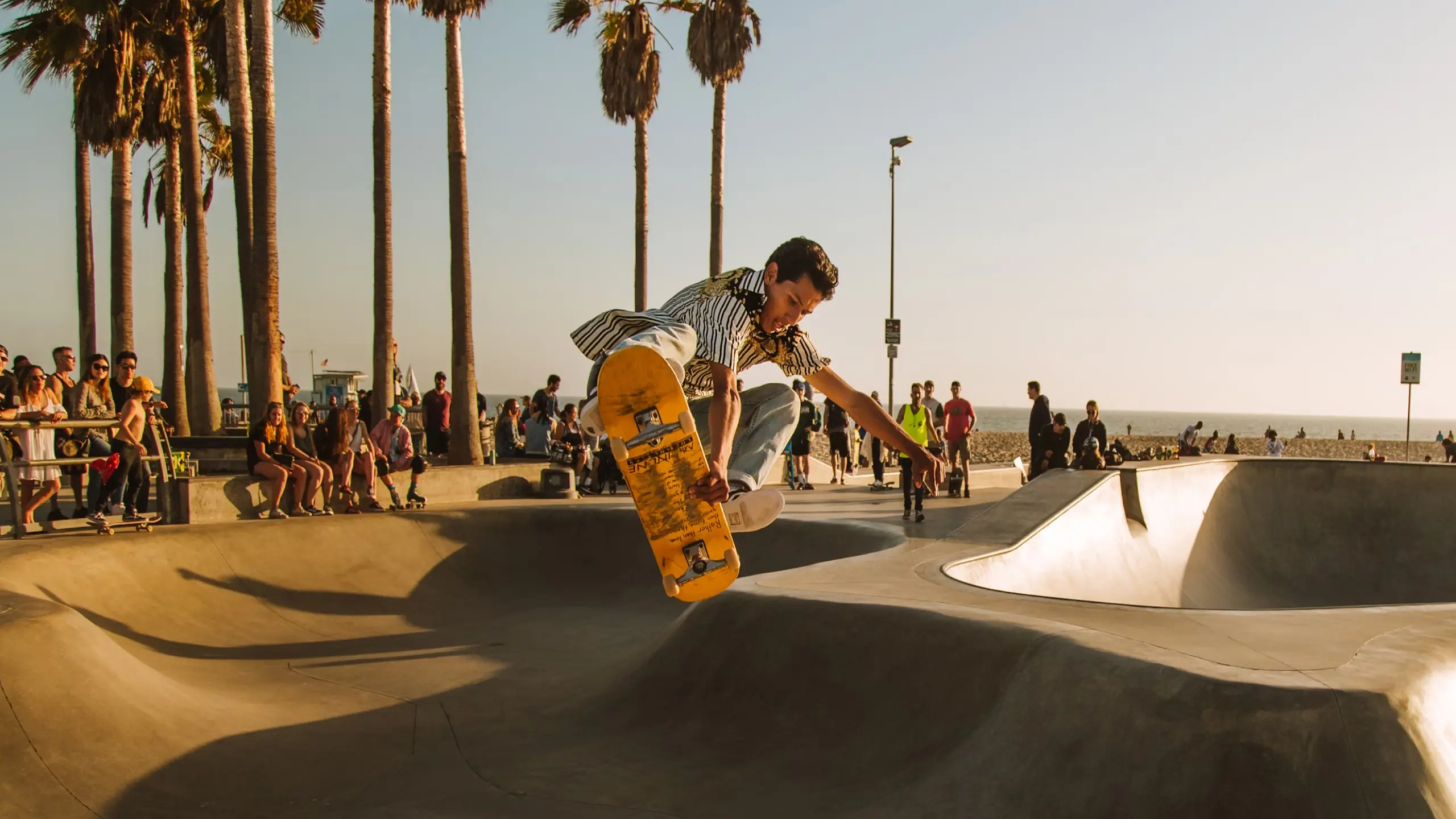 A skateboarder performing an aerial trick in a skate park near the beach, surrounded by onlookers and palm trees during sunset.