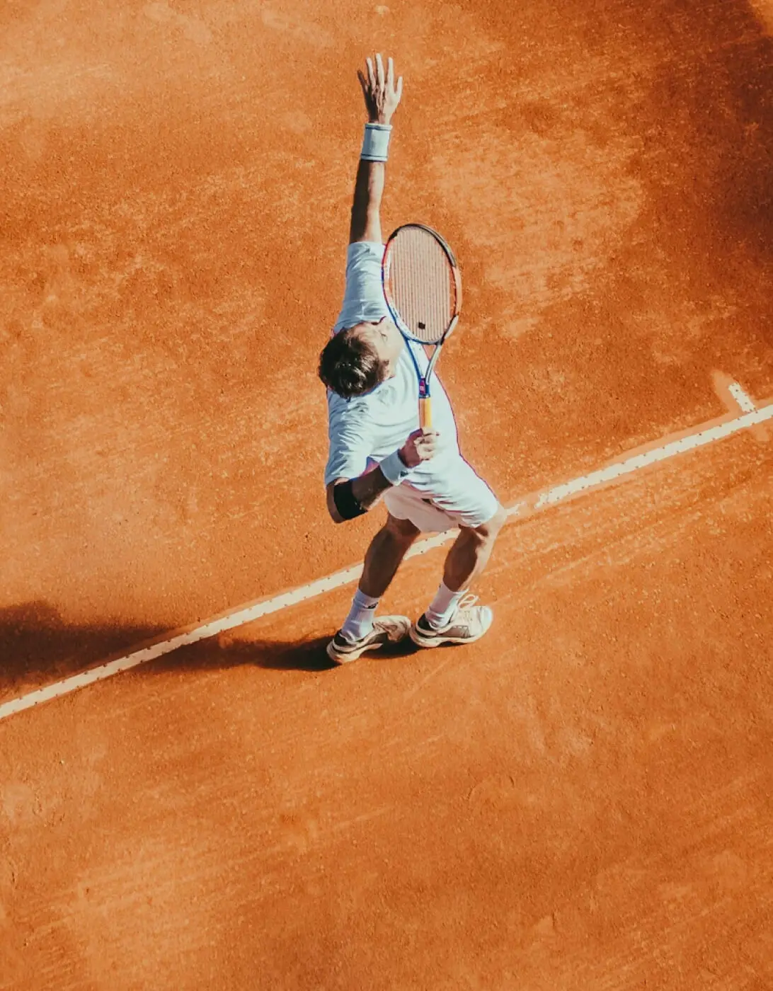 A tennis player in mid-serve on a clay court, captured from above, wearing a white outfit and raising their racket high in preparation to hit the ball.