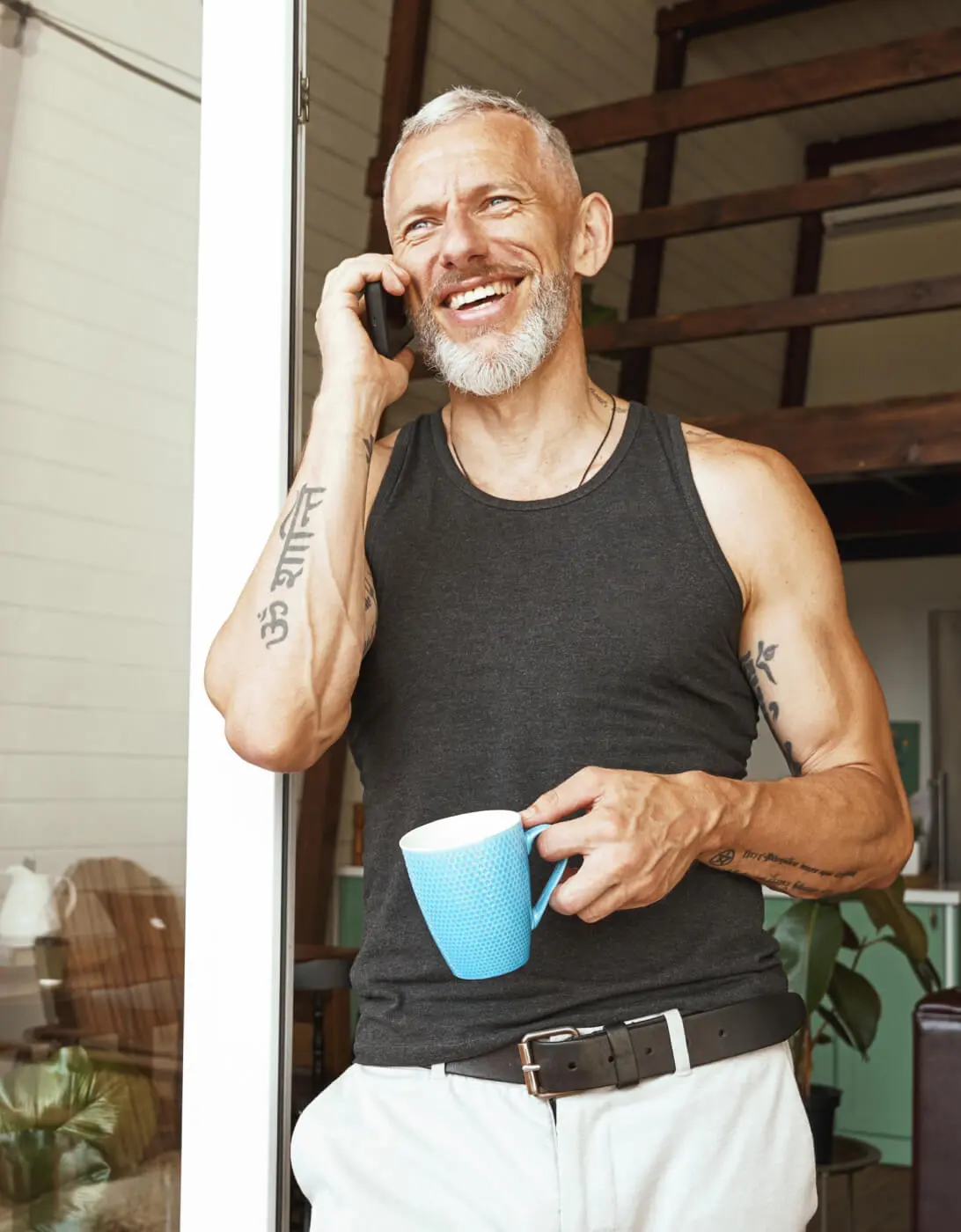 A smiling man with tattoos, holding a mug and talking on the phone.